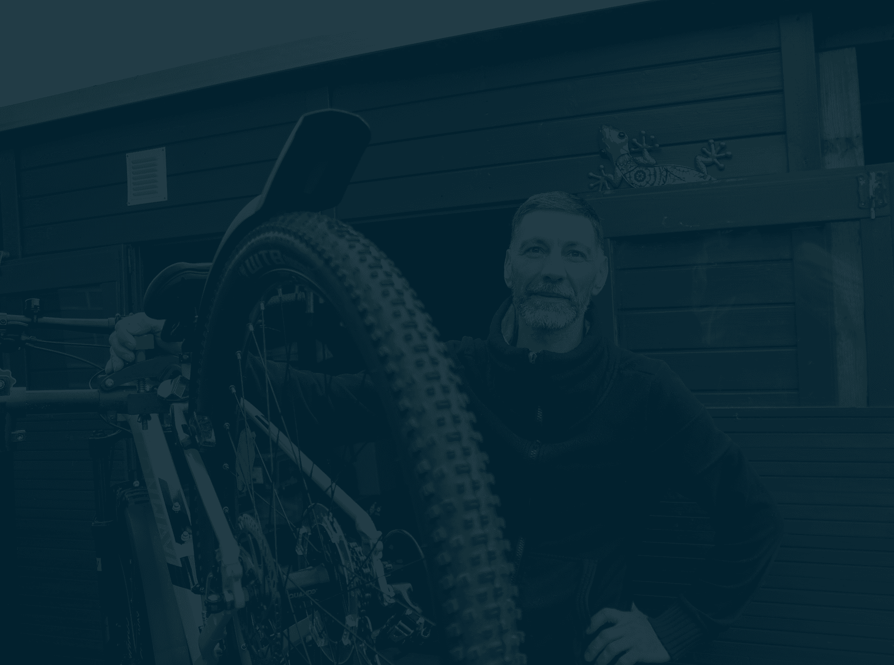 Ein bärtiger Mann steht lächelnd neben seinem Mountainbike vor einem dunklen Holzschuppen im Freien.