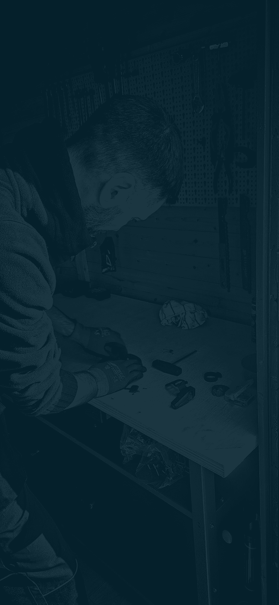 Ein Mann arbeitet konzentriert an einer Werkbank in einer Werkstatt mit Werkzeugen im Hintergrund.