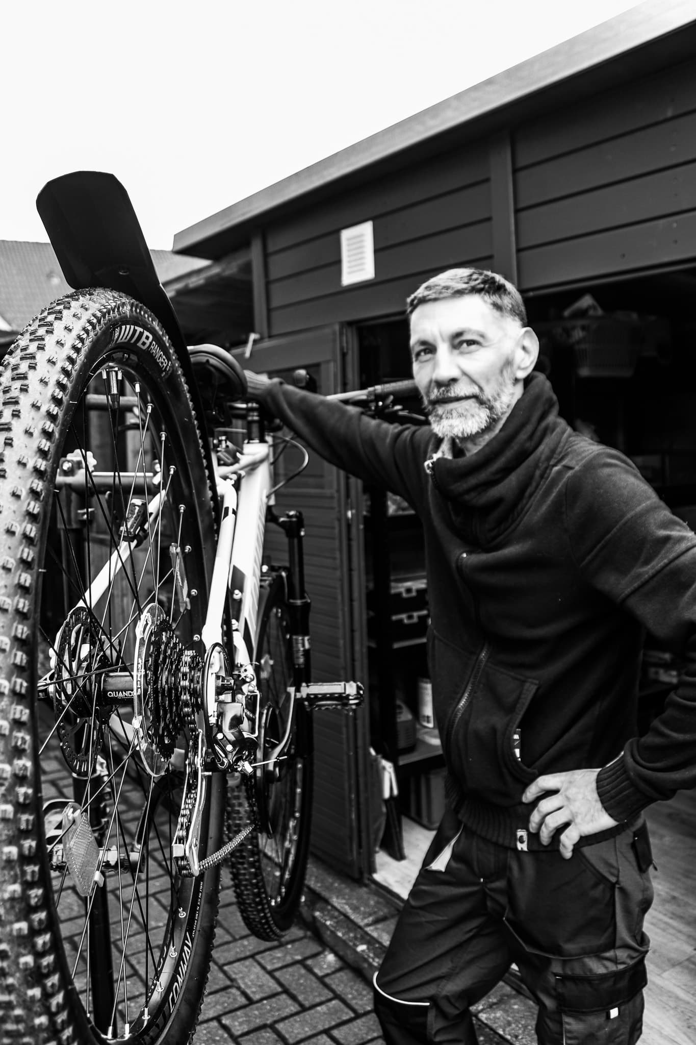 Ein bärtiger Mann posiert neben einem Mountainbike vor einem dunklen Holzschuppen in Schwarz-Weiß.