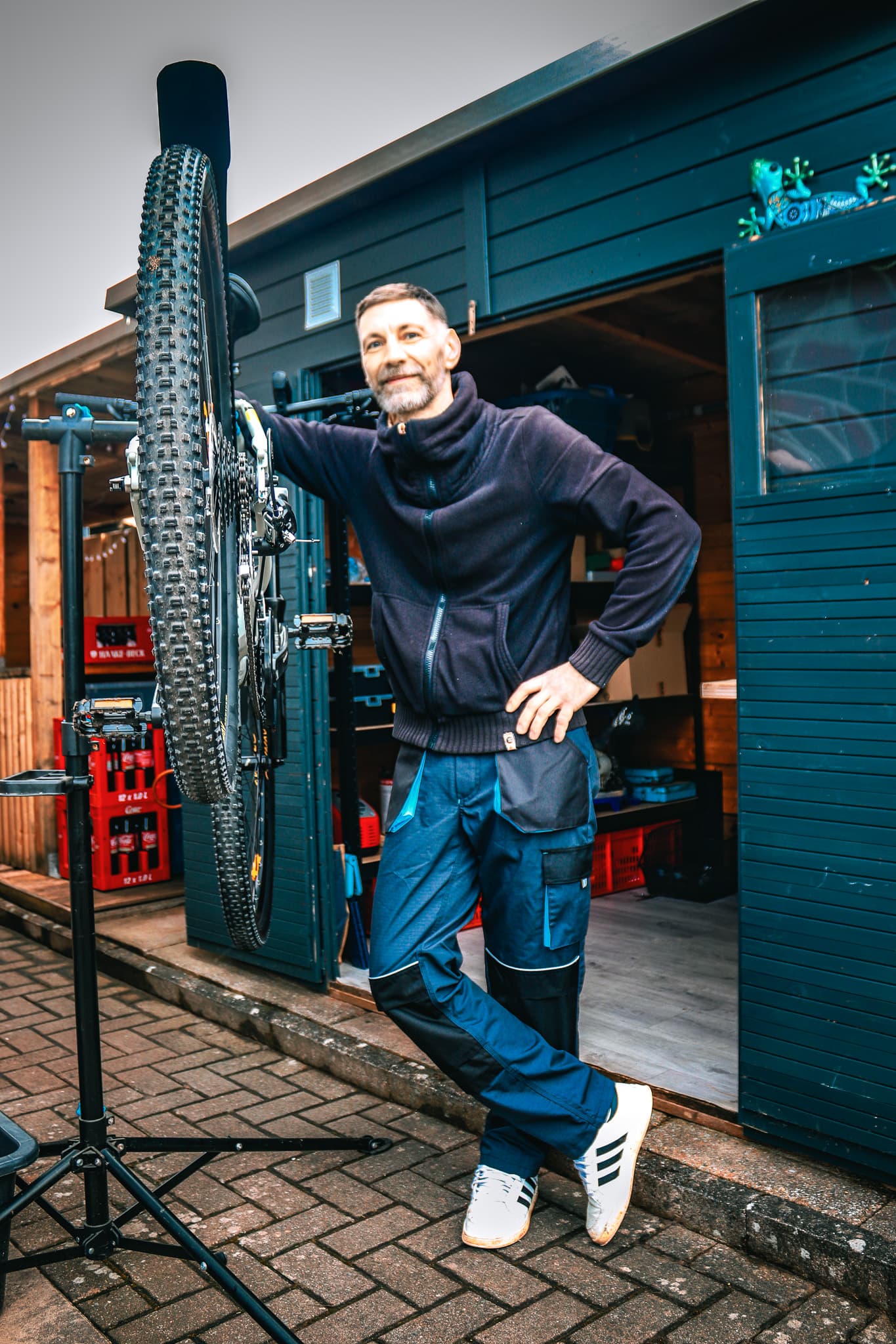Mann in blauer Arbeitshose steht neben einem vertikal montierten Mountainbike vor einer Gartenhütte.